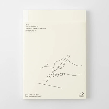 A cream-colored notebook or journal cover with minimalist black line art showing a hand writing '2027' on a pencil. The cover features Japanese text at the top left and a small 'MD' logo at the bottom right, set against a clean light gray background.