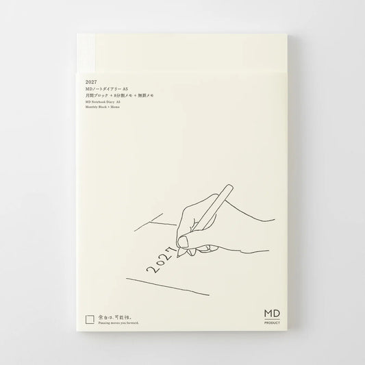 A cream-colored notebook or journal cover with minimalist black line art showing a hand writing '2027' on a pencil. The cover features Japanese text at the top left and a small 'MD' logo at the bottom right, set against a clean light gray background.